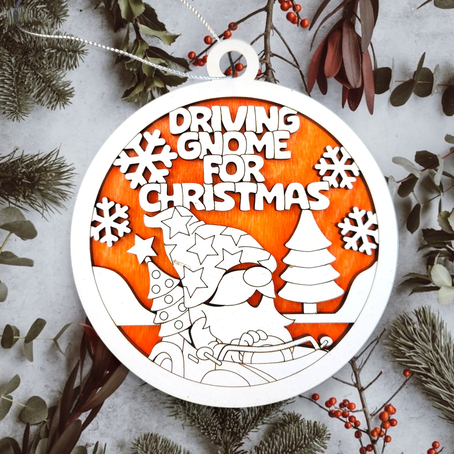 A circular Christmas ornament featuring a cartoon gnome driving a sled with a Christmas tree and snowflakes in the background.